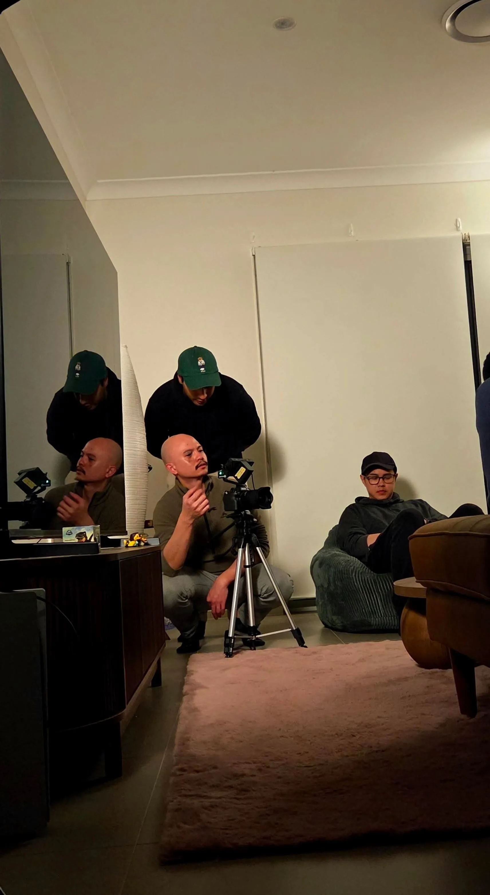 An actor crouching beside a camera on a tripod while the crew sets up a shot in a living room.